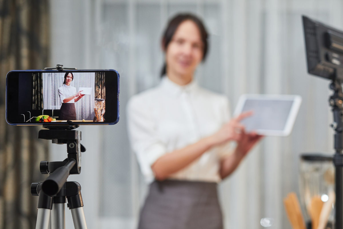 A woman holding an iPad stands in front of her cell phone, which is video recording on a tripod