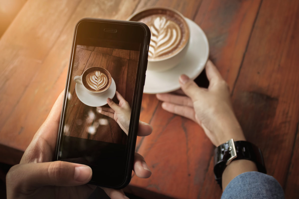 An image showing a person holding up there phone to take a photo of their latte for social media posting