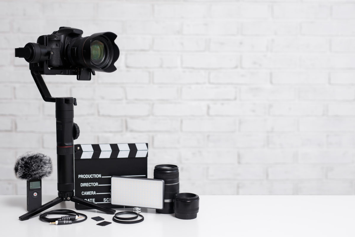 A production concept image featuring a camera, microphone, a clapperboard, and more in front a white brick wall.